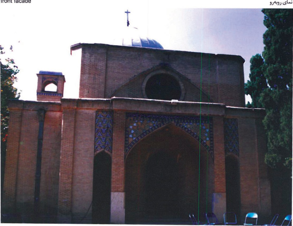 Front facade of the church — geometric brick volume with a small bell-cote dormer and pointed-arch entrance set behind a row of pencil cypress trees