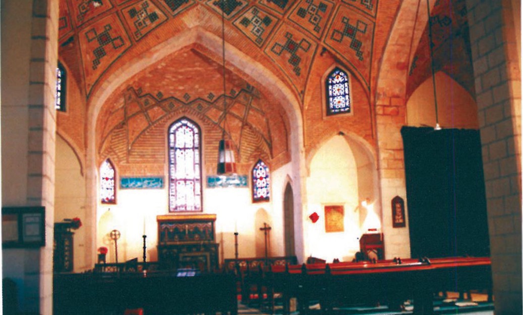 Interior of the church nave — pointed-arch arcade carrying a vaulted painted ceiling, lantern hanging at the centre with rows of wooden pews and stained-glass window beyond