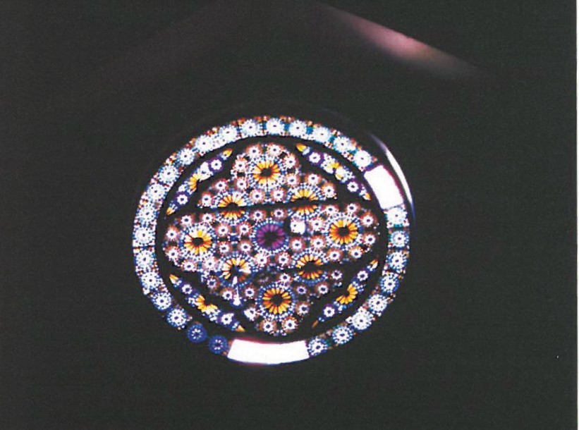 Round rose window above the church entrance — radiating geometry of stained-glass petals filled with red, blue and green floral motifs