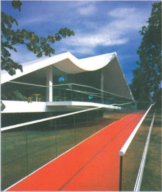 Curved white-canopy summer pavilion in Kensington Gardens — billowing fabric roof above a glass-walled exhibition space, with a long red carpet path leading to the entrance