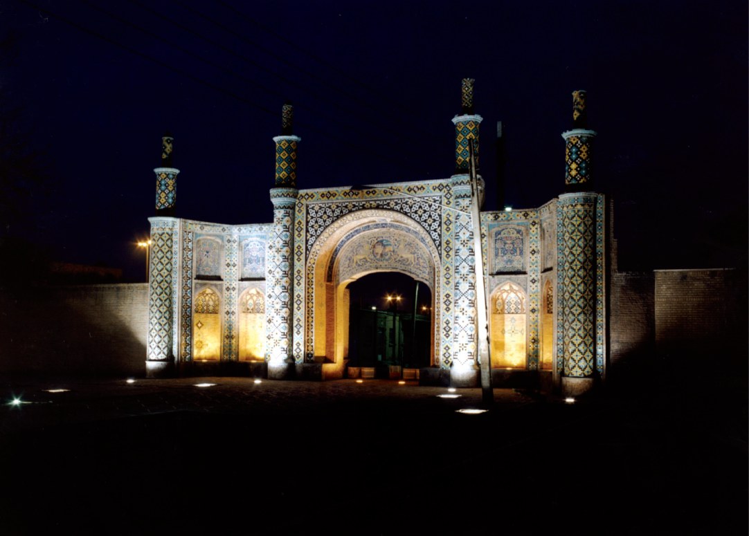 A Qajar-era tiled portal with four minaret-like turrets, dramatically illuminated at night by ground-level light fixtures