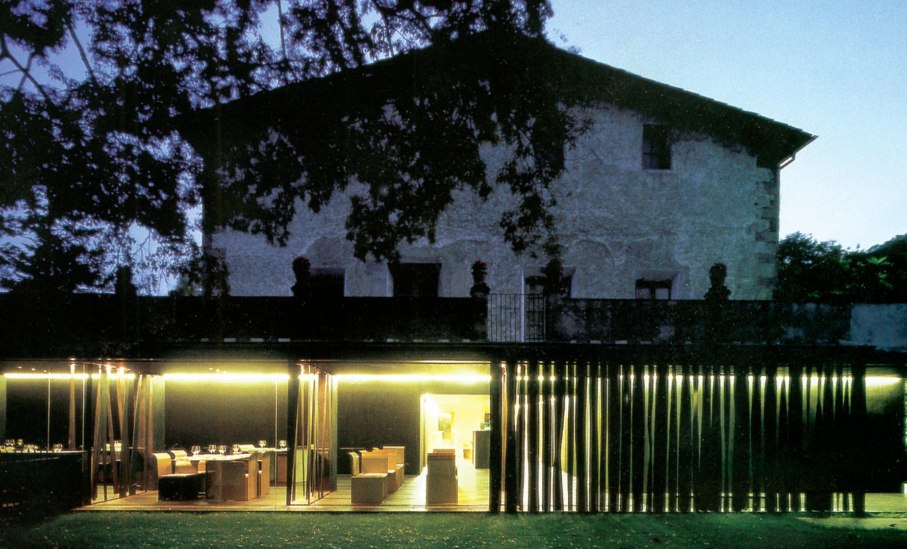 Night exterior view of Les Cols showing the lit-up steel-and-glass pavilion housing the dining room beneath the dark stone masia