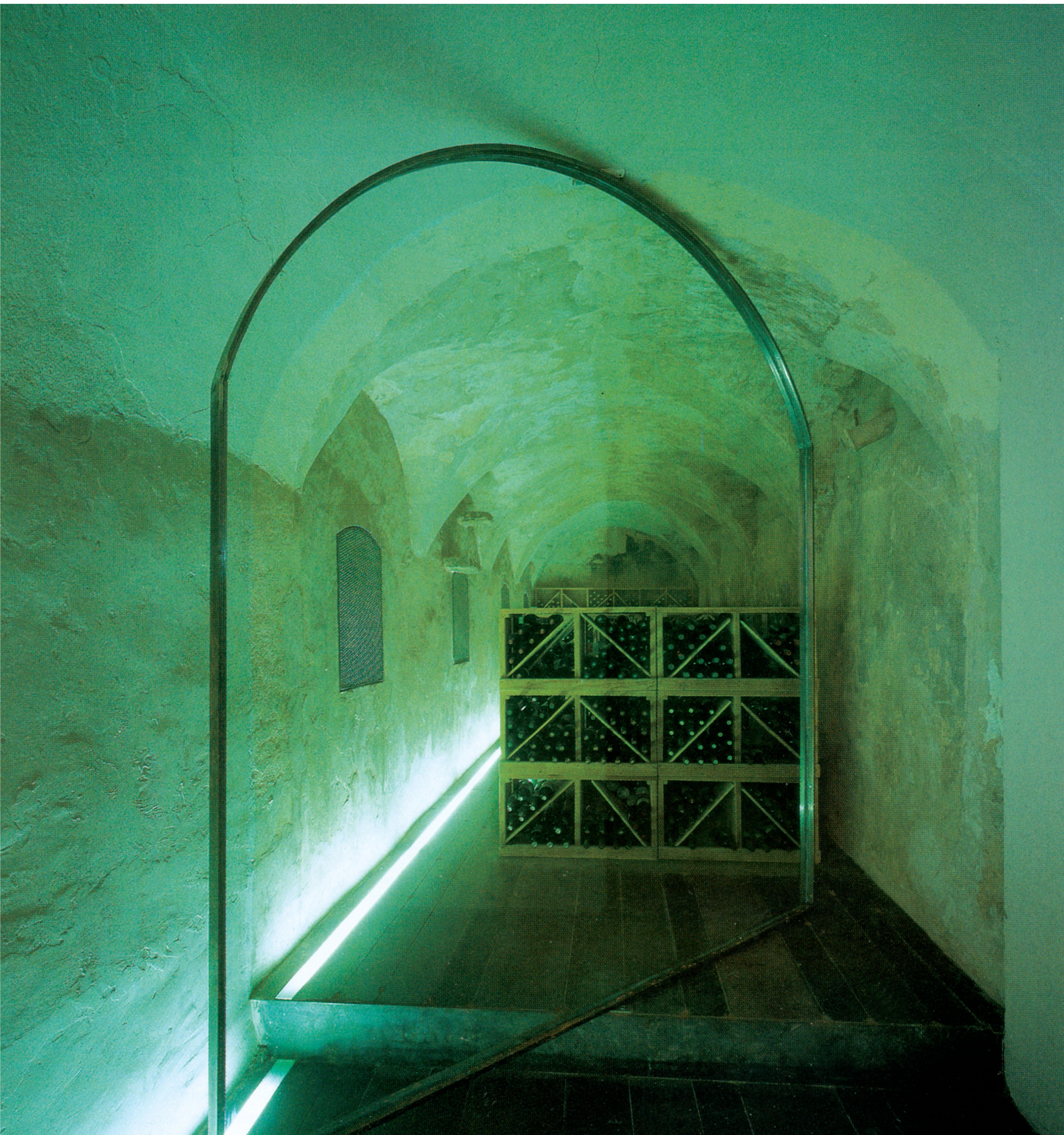 Wine cellar set into the original building's stone vaults, with a simple glass door, a strip of light embedded in the floor, and wooden bottle racks beyond