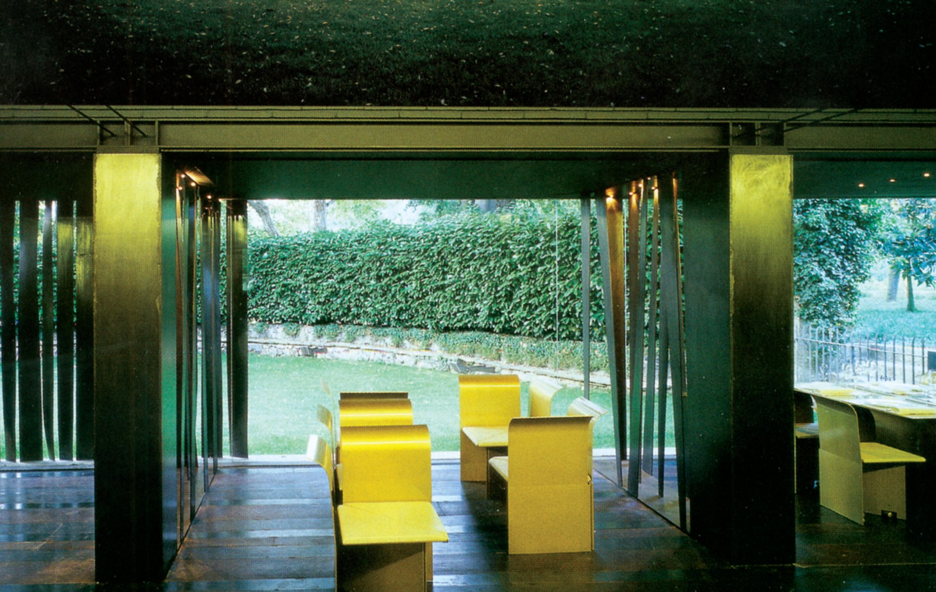 Interior of the main dining room, with yellow furniture and steel screens opening through frameless glass onto a lush garden