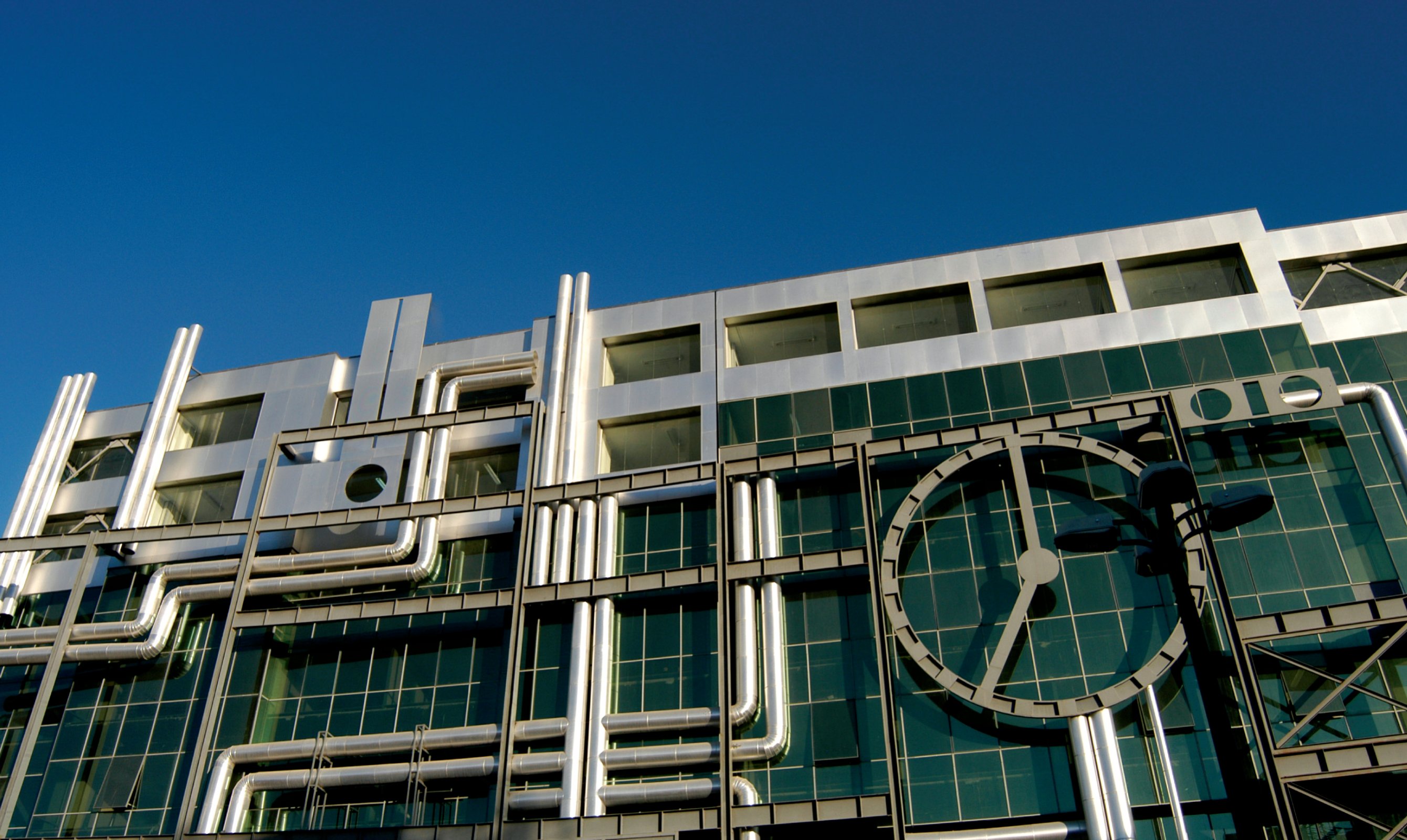 Detail of the Gas Company facade — polished stainless-steel pipes laid across a glass curtain wall, with a large mechanical clock motif at the right