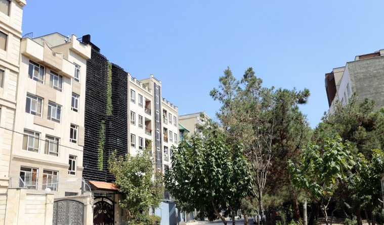 نگاه از آن سوی خیابان: دیوارِ میخ‌دار به‌مثابه یک پیکره تیره میان آپارتمان‌های پیرامون خوانده می‌شود، و نوار سبز کاشته‌شده از زمین تا بام امتداد می‌یابد.