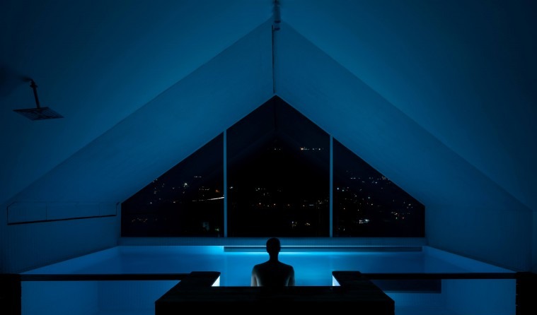 Pool at night with man's silhouette standing by the triangular gable window overlooking dark city lights