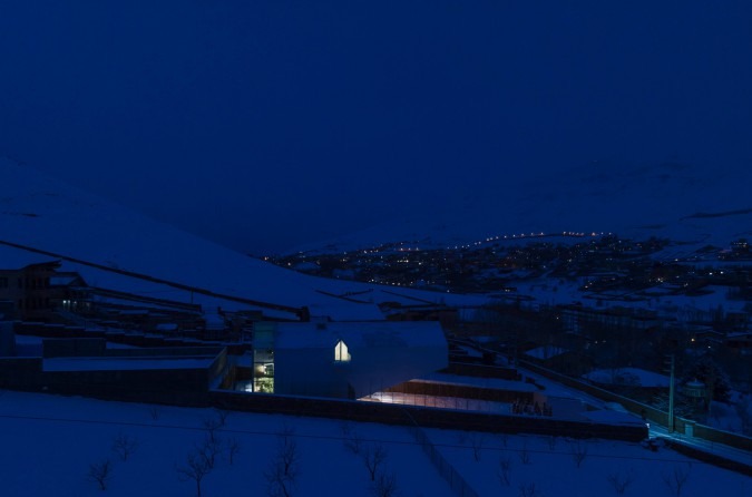 Snowy night with city lights below the snowy hills and the lit interior of the gable