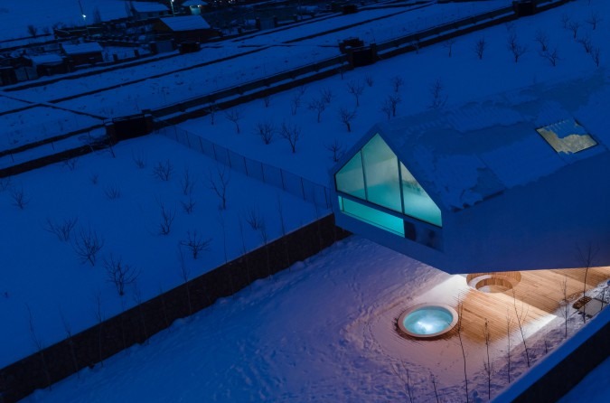 Snow night exterior with the glowing teal triangular pool window emerging from the white volume