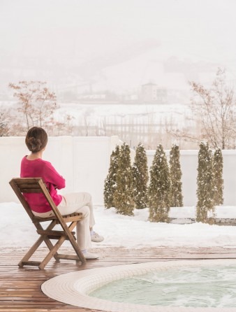 Snow patio with woman seated on a wooden chair facing the misty mountain view with jacuzzi in the foreground