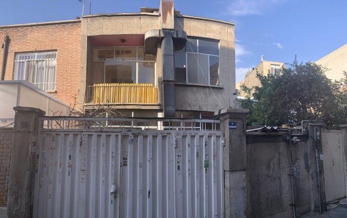 چهره‌ی خیابانیِ پیش از کار، از کوچه: یک دروازه‌ی فلزی با ناودانیِ ورقی، آجرِ پیرِ طبقه‌ی اول و یک پنجره با نرده‌ی زرد ـــ همان مقیاسِ کوچه که پروژه نشانه‌های خود را از آن می‌گیرد.
