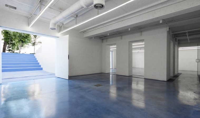 Nabshi Gallery's main ground-floor exhibition room with a blue resin floor and a wall opening onto the blue courtyard