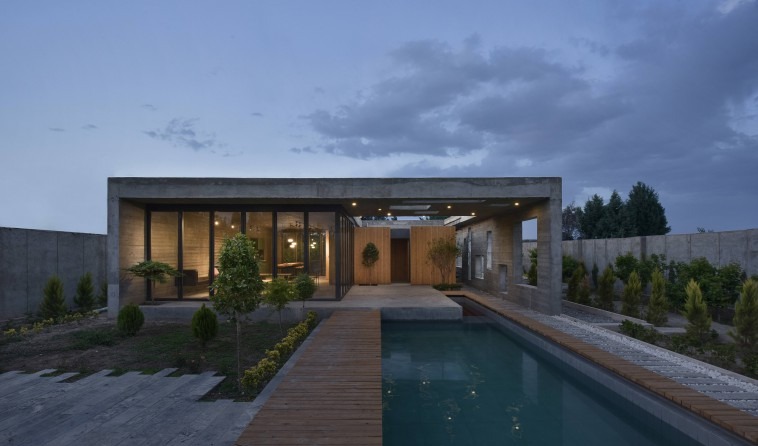 Twilight frontal view of the project: the concrete frame reads as a single horizontal mass, the long timber deck and poo