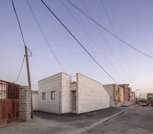 Twilight street view of the white-stone clad house showing the cubic mass and arched vault behind, in the dense Najafabad fabric